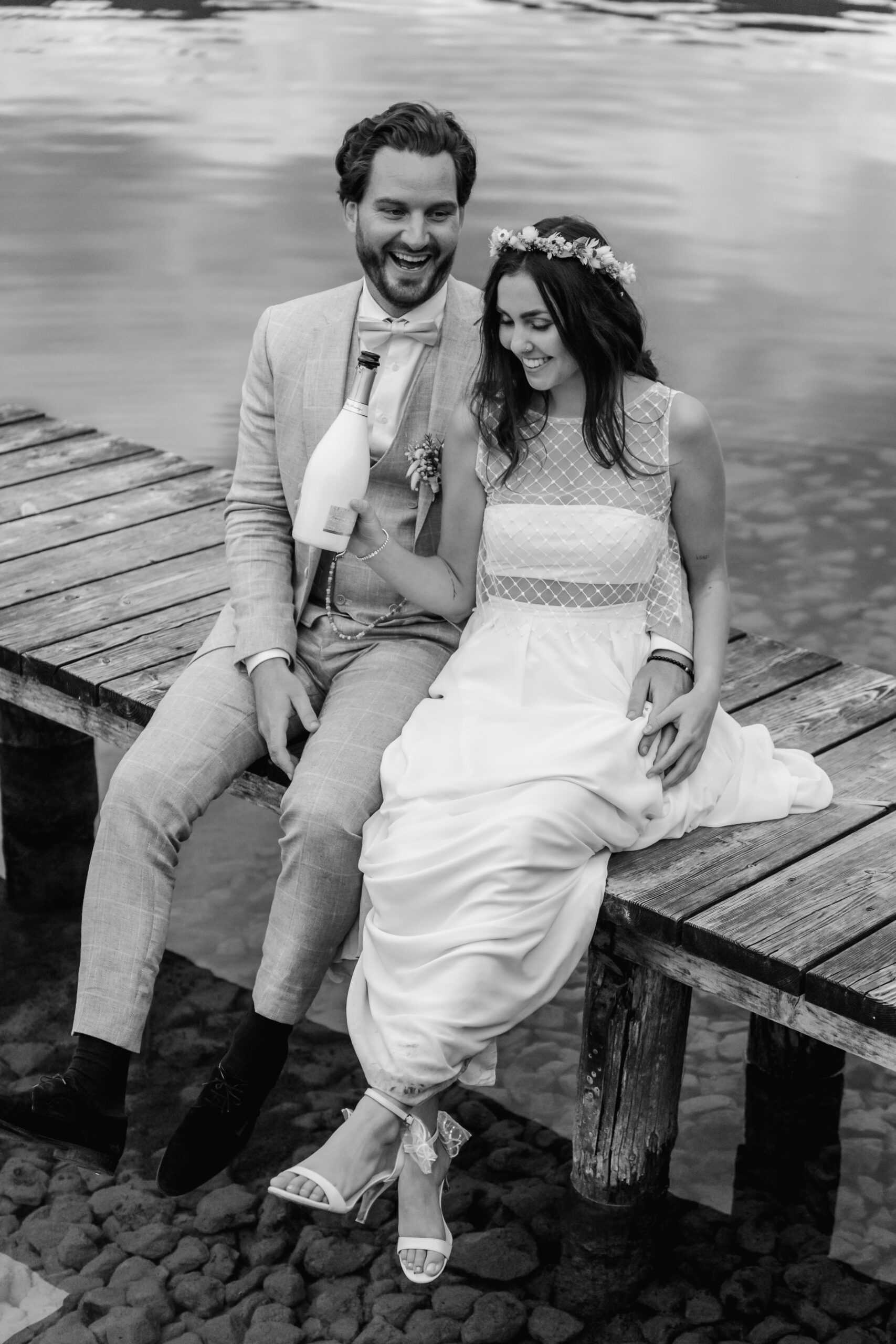 Glückliche Braut und Bräutigam sitzen auf dem Steg am Tegernsee und trinken Champagner