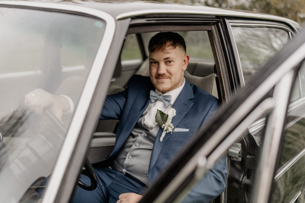 Bräutigam sitzt im Auto, lächelt und schaut direkt in die Kamera, Hochzeitsfotograf Frankfurt