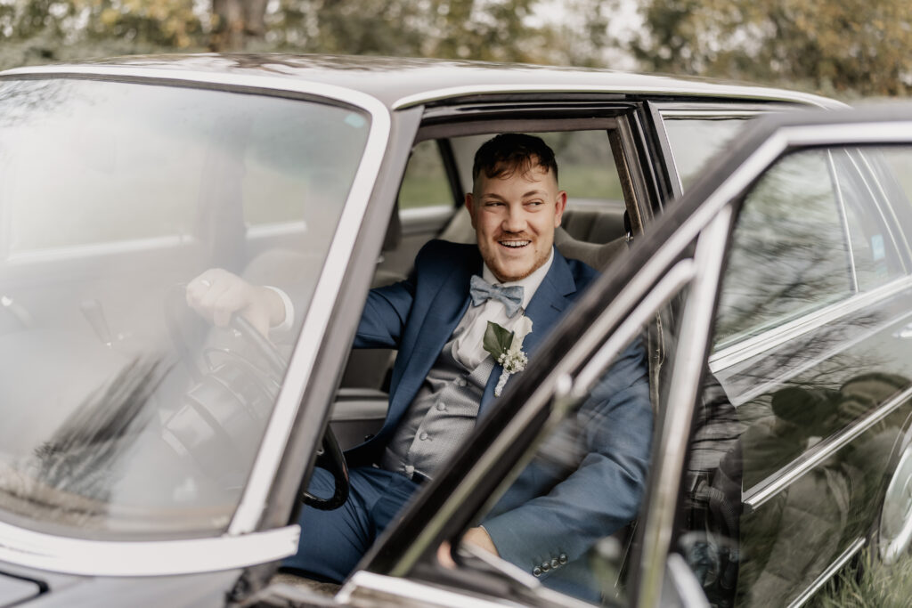 Bräutigam sitzt lachend im Oldtimer und schaut aus der offenen Tür, Hochzeitsfotograf Frankfurt