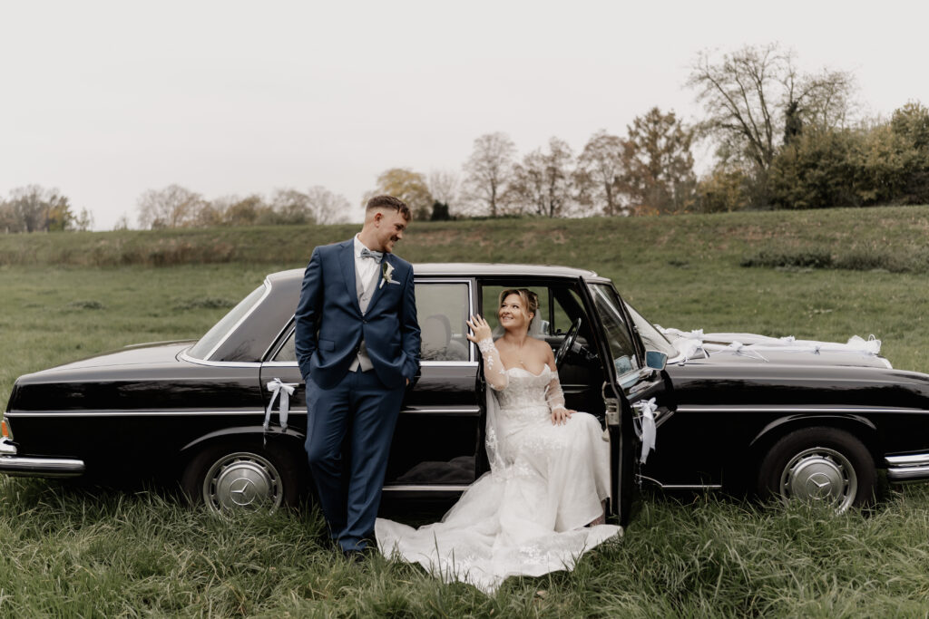 Braut sitzt in der Autotür und Bräutigam lehnt lächelnd am Auto. Hochzeit Mönchshof Eddersheim