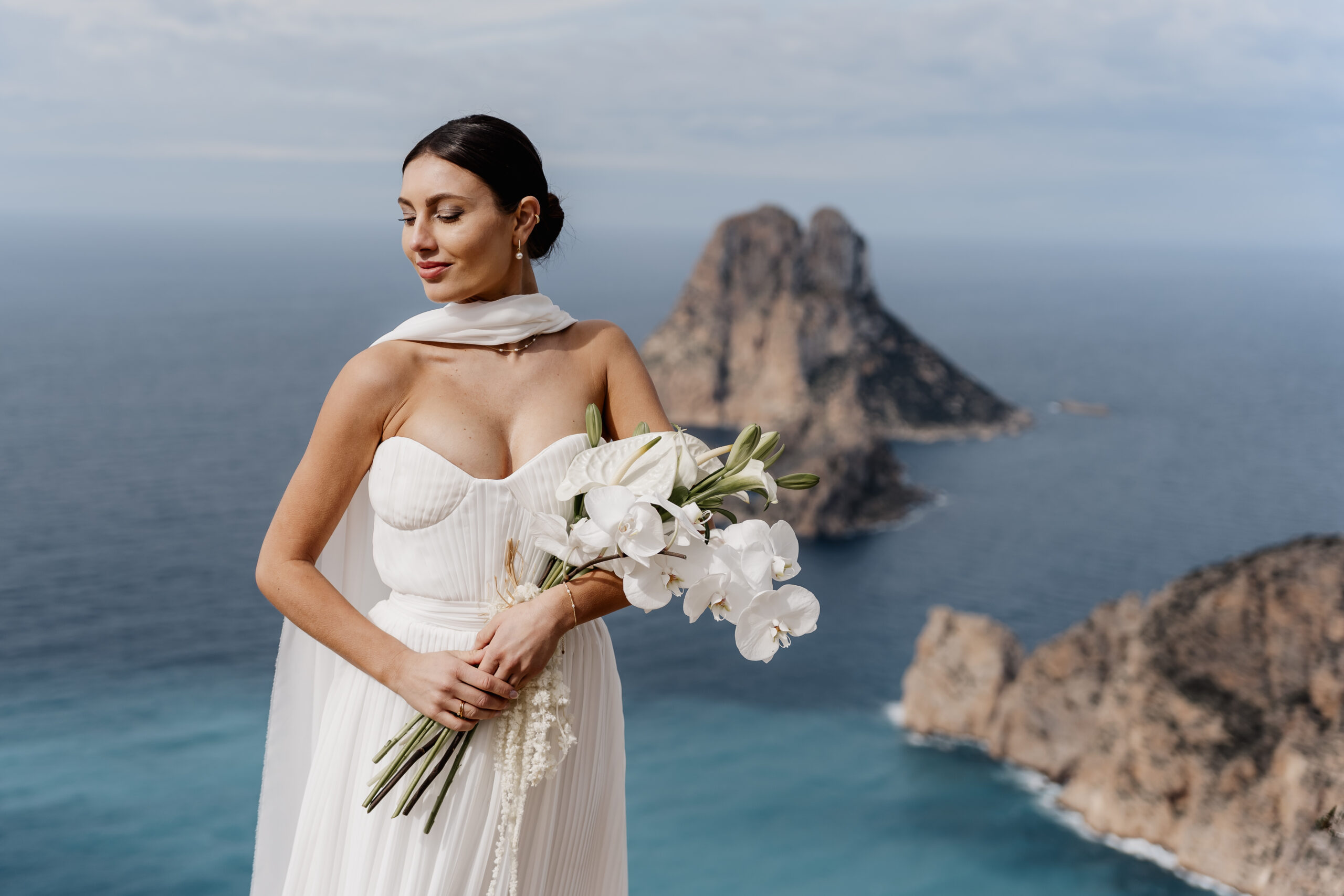 Elegante Braut steht vor dem Es Vedra auf Ibiza und hält ihren Brautstrauß
