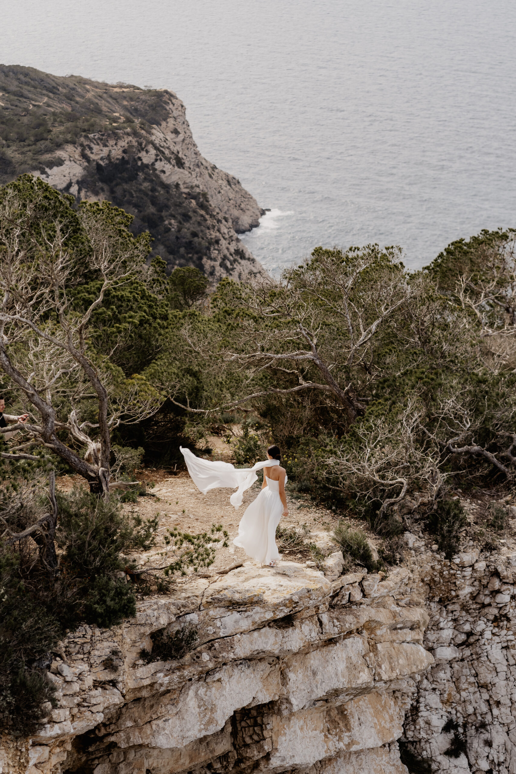 Braut steht mit wehendem Schleier auf einer Klippe von Ibizas Küste, Hochzeitsfotograf Ibiza