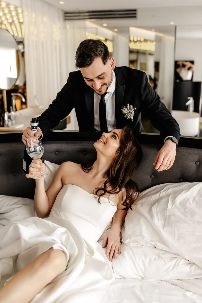 Locker-liebevoller Moment im Bett – Brautpaar genießt Sekt im Roomers Hotel München