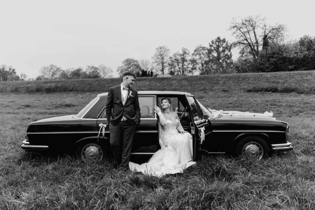 Schwarz-Weiß-Foto,Braut sitzt in der Autotür, Bräutigam lehnt am Wagen, Hochzeit Mönchshof Eddersheim