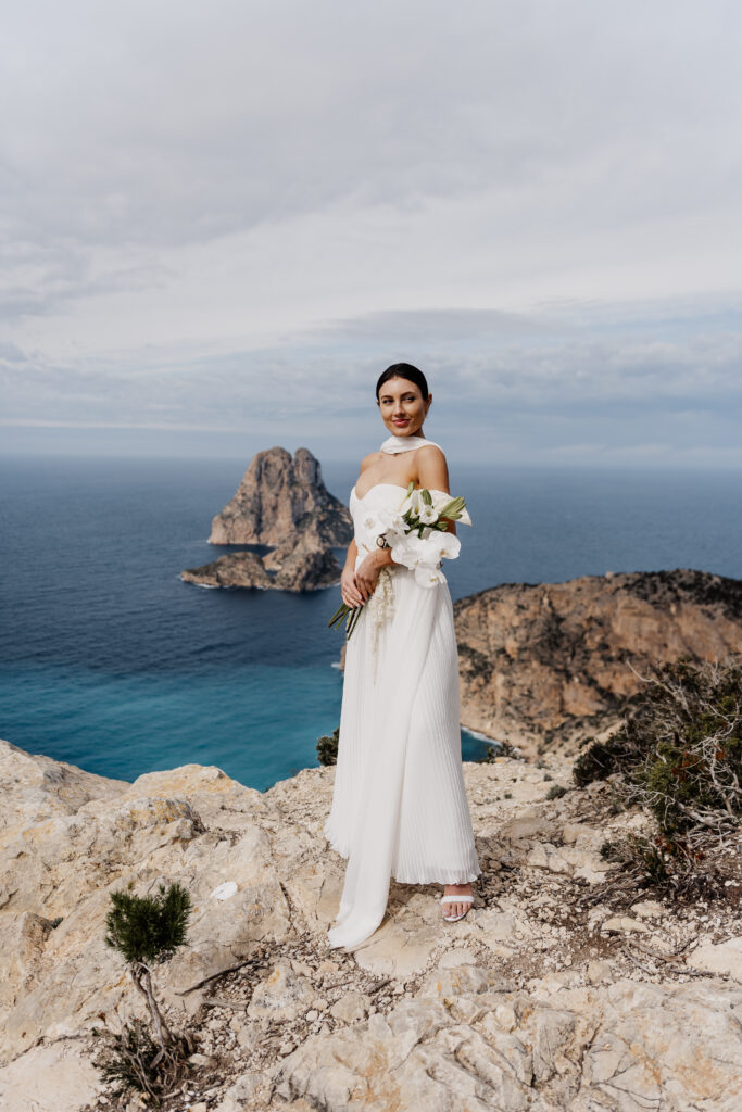 Stolze Braut mit weißem Kleid und Blumen, Blick auf den es Vedra Ibiza, Elopement photographer Ibiza