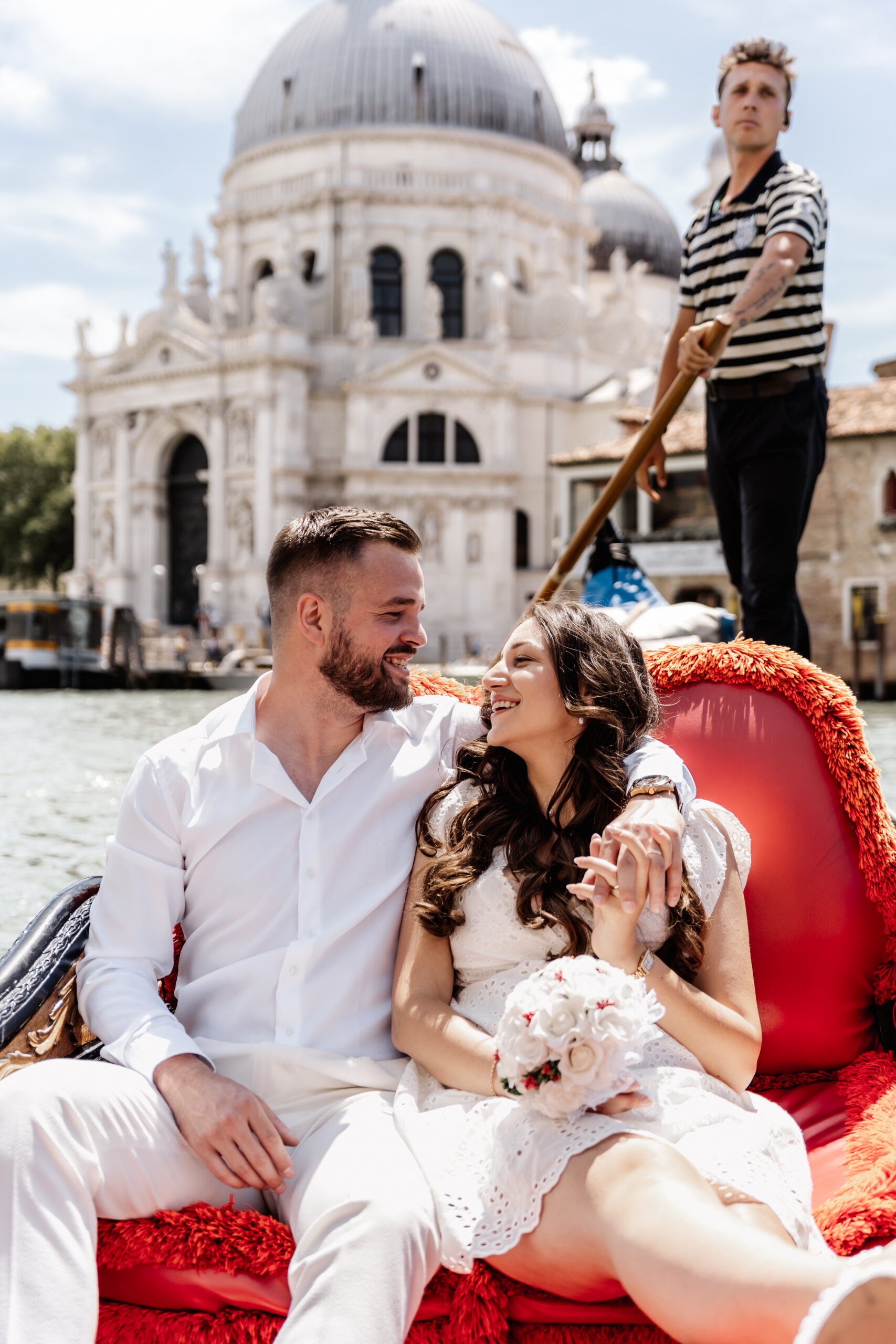 Braut und Bräutigam sitzen eng umschlungen in einer Gondel, Gondolier schaut in die Ferne, Sehenswürdigkeit im Hintergrund