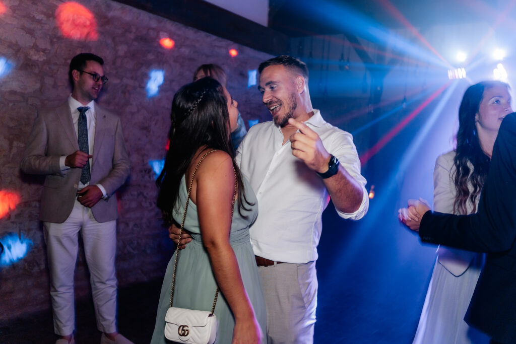 Paar tanzt lachend auf der Hochzeitsparty, farbiges Licht und Gäste im Hintergrund