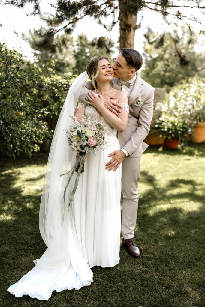 Brautpaar umarmt sich im Grünen, Bräutigam küsst die Braut, Hochzeit im BourdierKoeller Stetten