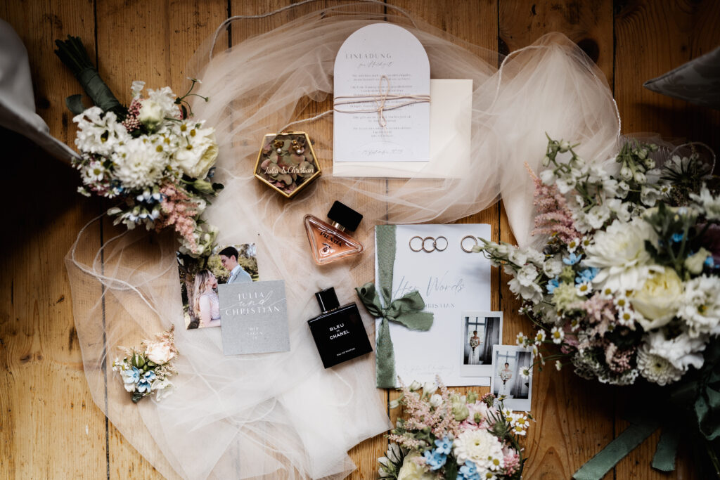 Flatlay mit Hochzeitseinladung, Schleier, Ringen und Blumen, stilvolle Hochzeitsdetails auf Holzboden