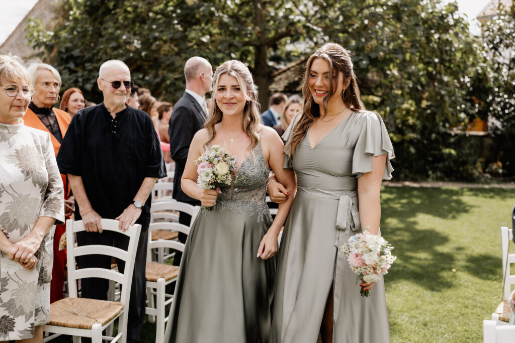 Brautjungfern gehen lächelnd zum Trauplatz, Hochzeitsgäste stehen im Garten der Eventlocation