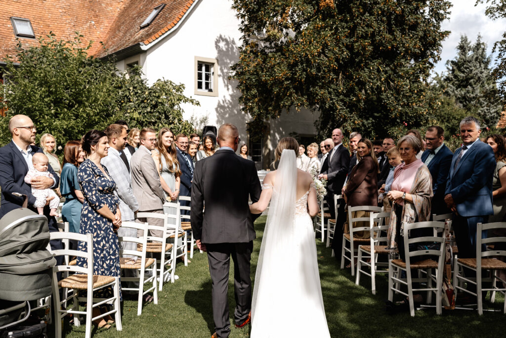 Braut und Vater gehen den Mittelgang entlang, Hochzeitsgäste stehen links und rechts