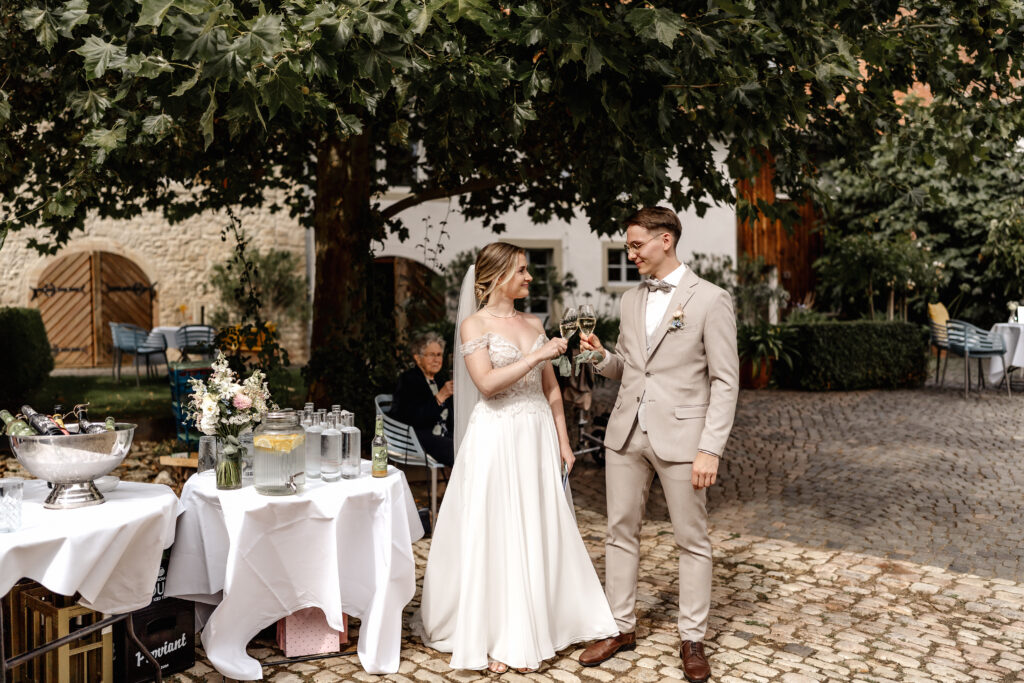 Brautpaar stößt mit Champagnergläsern beim Sektempfang unter Bäumen an