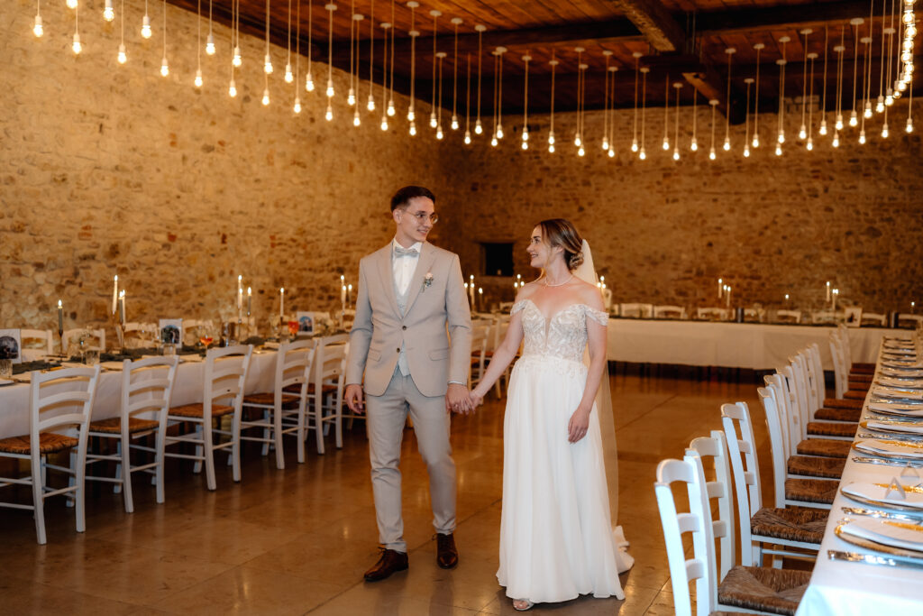 Brautpaar hält sich an den Händen und geht durch den festlich gedeckten Saal unter warmen Lichterketten