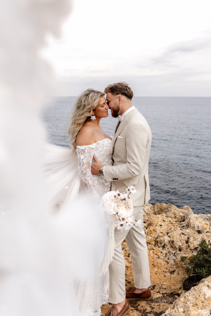 Hochzeitspaar kuschelt innig an Mallorcas Küste mit wehendem Brautschleier